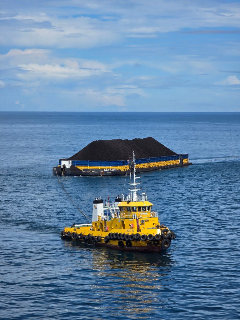 A yellow tugboat towing a coal barge across the calm blue sea under a partly cloudy sky.